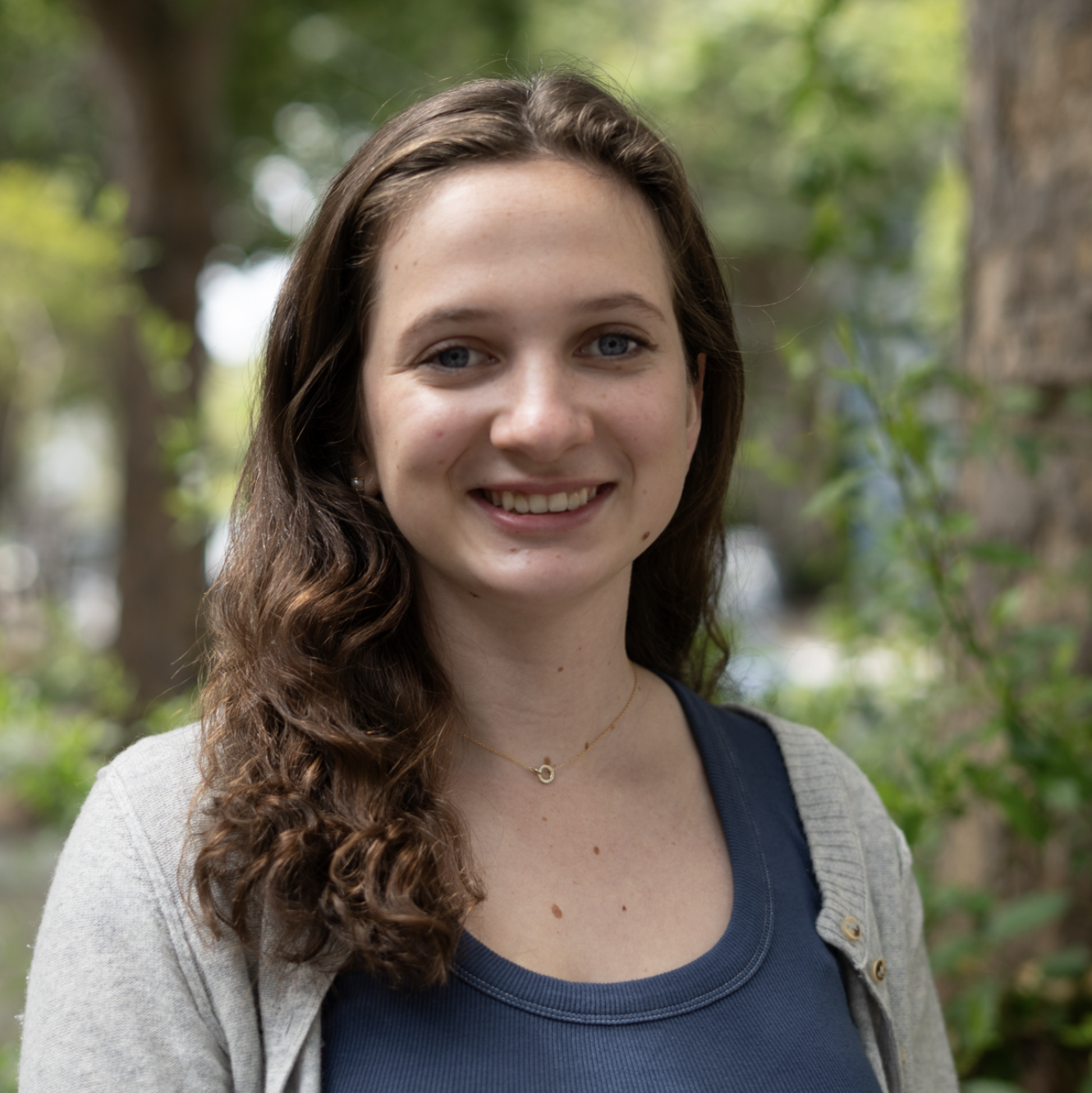 A young woman with long brown hair and a blue top smiles outdoors, with trees and greenery in the blurred background.